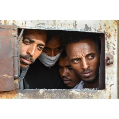 Men look out of an opening in the main gate of a warehouse in Zintan detention center, south of Tripoli, where 700 people are being detained. LIBYA 2019 © Jérôme Tubiana/MSF
