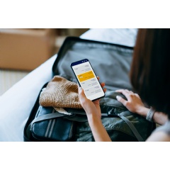 Over the shoulder view of young Asian woman using smartphone, checking digital flight ticket and boarding pass on device screen while packing a suitcase on bed for a trip. Planning for travel. Travel and vacation concept